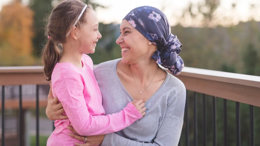 A mother and cancer survivor is sitting, with one arm around her daughter.