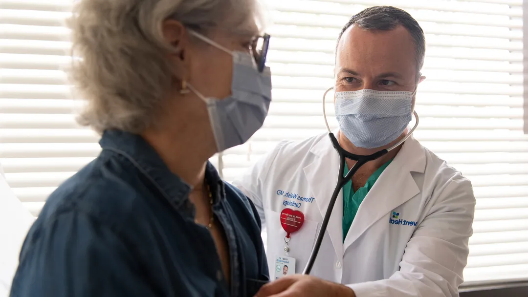 Doctor listening to patient's heartbeat with a stethoscope while both wear masks.