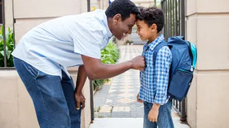A father and a son standing outside,. The son is wearing a backpack and the father is bending over, comforting him.