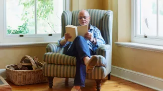 An older man reading a book while seating on a chair.