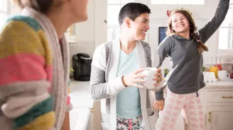 A woman dancing in the kitchen with her daughters and drinking coffee.