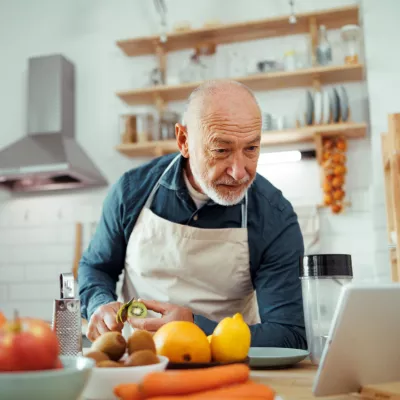 A Senior Reads A Recipe From a Tablet While Chopping Vegetables in His Kitchen