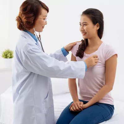 A woman is sitting on an exam table while a doctor listens to her heart.
