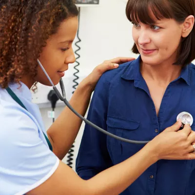 A woman sitting on an exam table, having her lungs listened to by a doctor.
