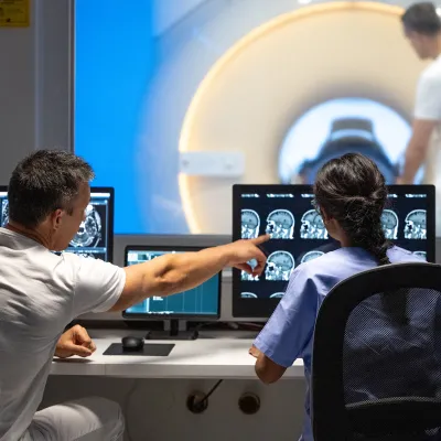 A male and female imaging technician looking at CT scans of a patient's head in the foreground while another male technician is prepping the patient in the CT machine in the background.