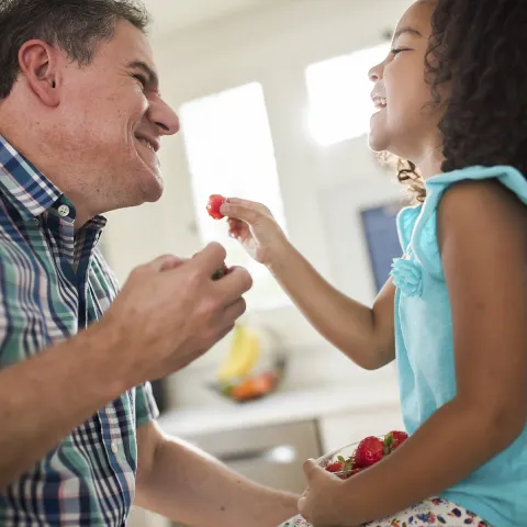 A grandfather and his granddaughter laugh as they eat strawberries in the kitchen.