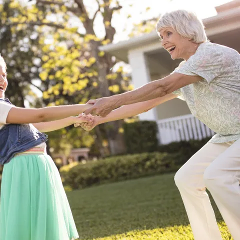 A Caucasian grandmother and granddaughter spin around and dance in a yard.