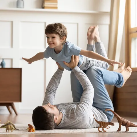 A Little Boy Plays "Air Play" on his dad's legs on the floor with dinosaur toys surrounding them