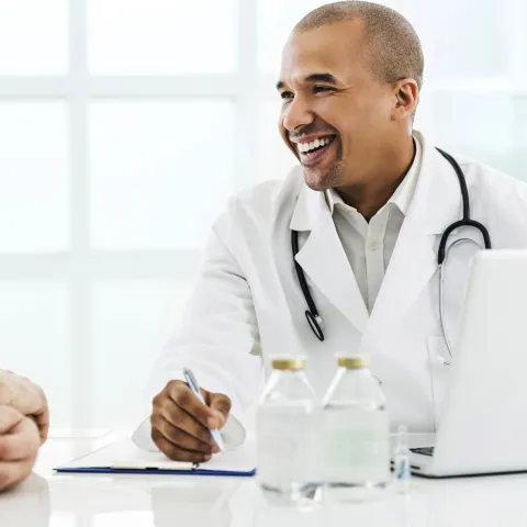 A senior man visits his male doctor for his annual screening. Both are seated at a table.