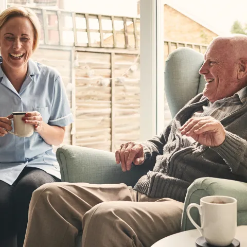A senior man, reclined in a chair, laughs with his caretaker, who is seated next to him.