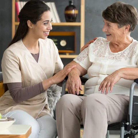 A woman in a wheelchair, with her caretaker sitting next to her and comforting her. Both are smiling.