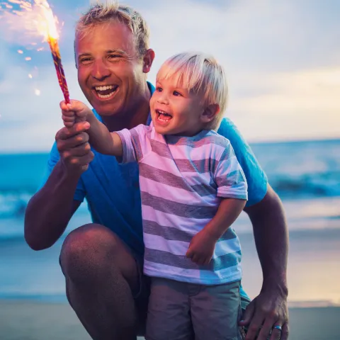 A father and son holding a firework on the beach at sunset.