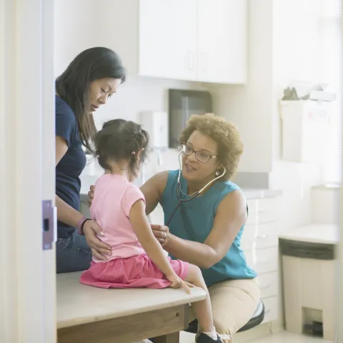 A female AHS doctor listens to a little girl's heart in the exam room while her mother comforts her