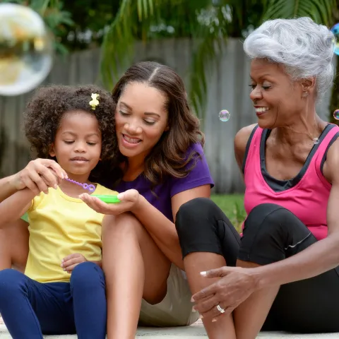 A grandmother, mother, and daughter play with bubbles outside. 
