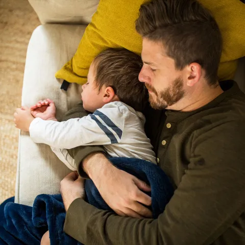A dad is embracing his son while they are both napping on the couch.
