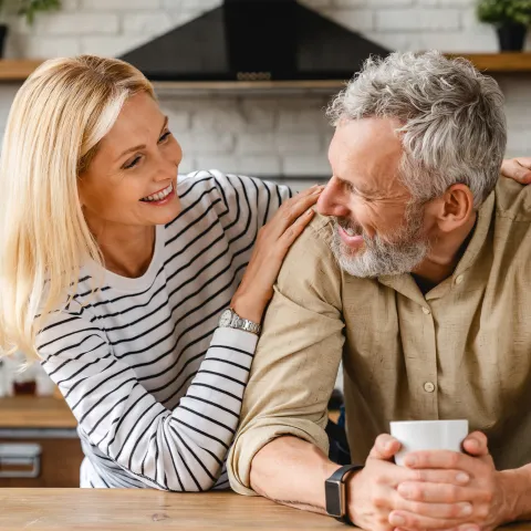 An older couple smiling at each other while in a kitchen.