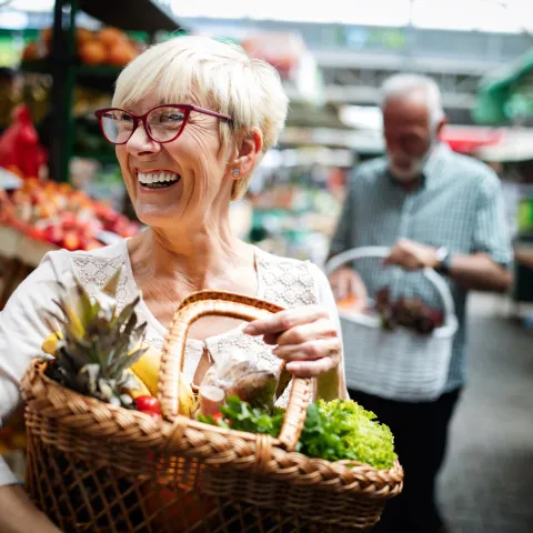Woman smiling in the foreground while her husband is in the background. Both are holding a basket of fresh produce at a farmers market.