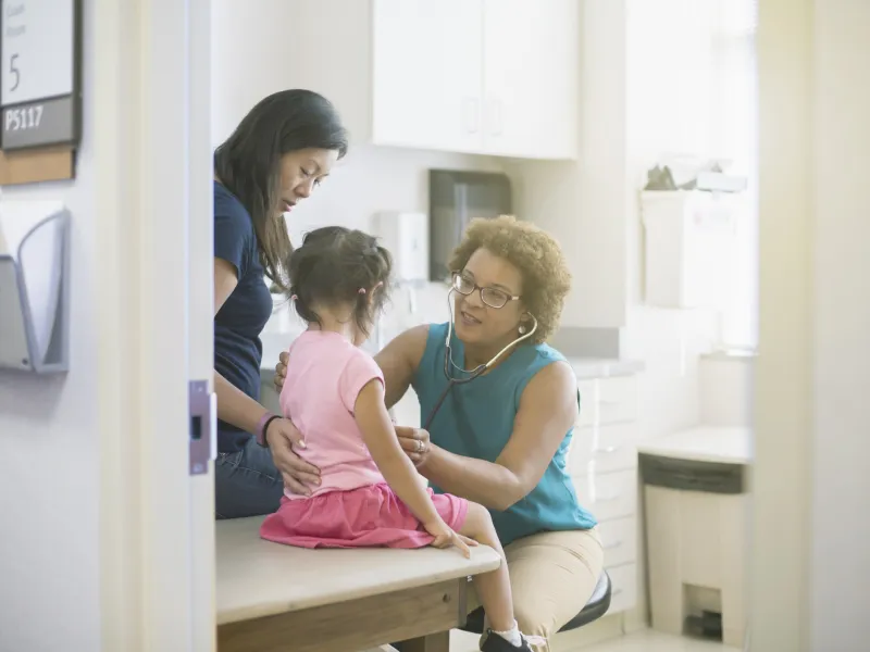 A female AHS doctor listens to a little girl's heart in the exam room while her mother comforts her