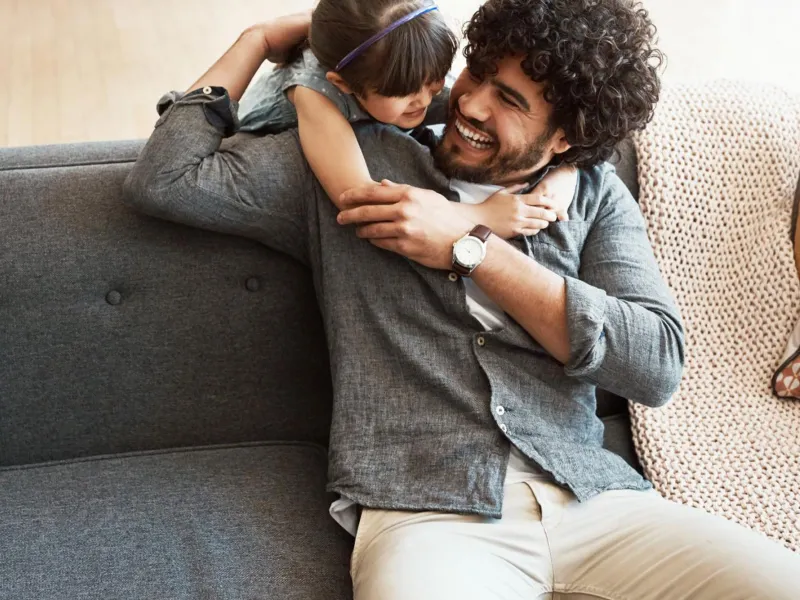 A daugther hugging her father who is sitting in the couch.