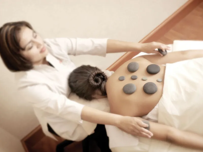 A woman laying down on a bed getting a warm stone massage done on her back.