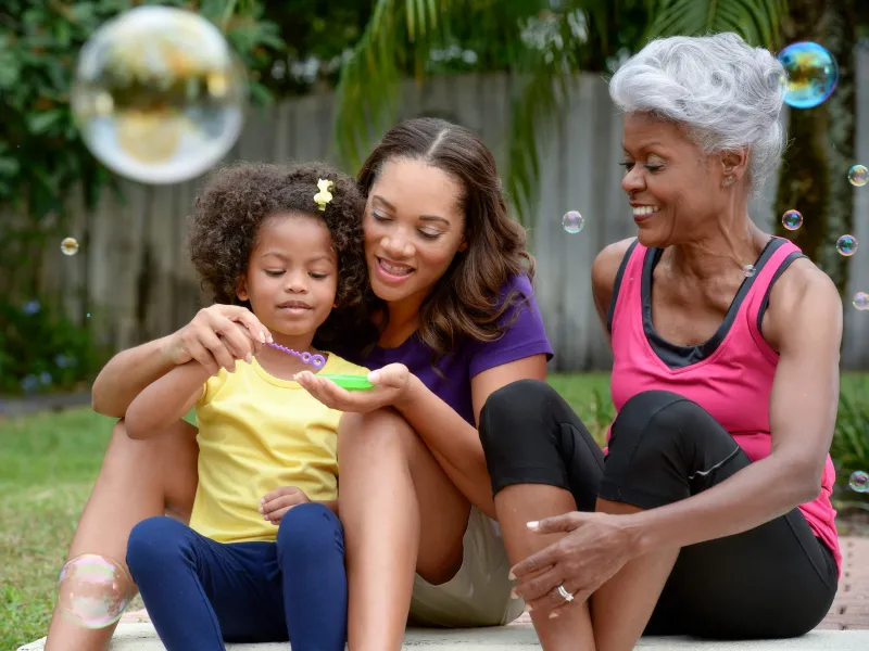 A grandmother, mother, and daughter play with bubbles outside. 