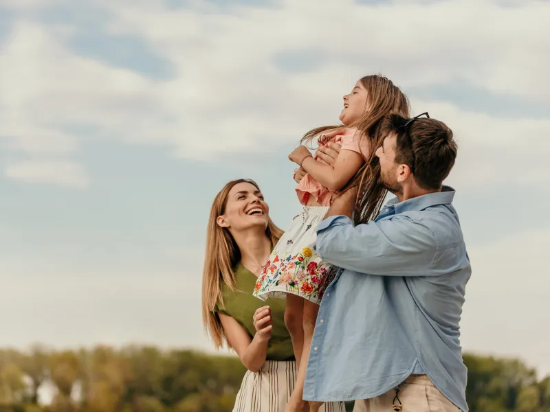 A family of a mother, father and daughter smiling while outdoors.