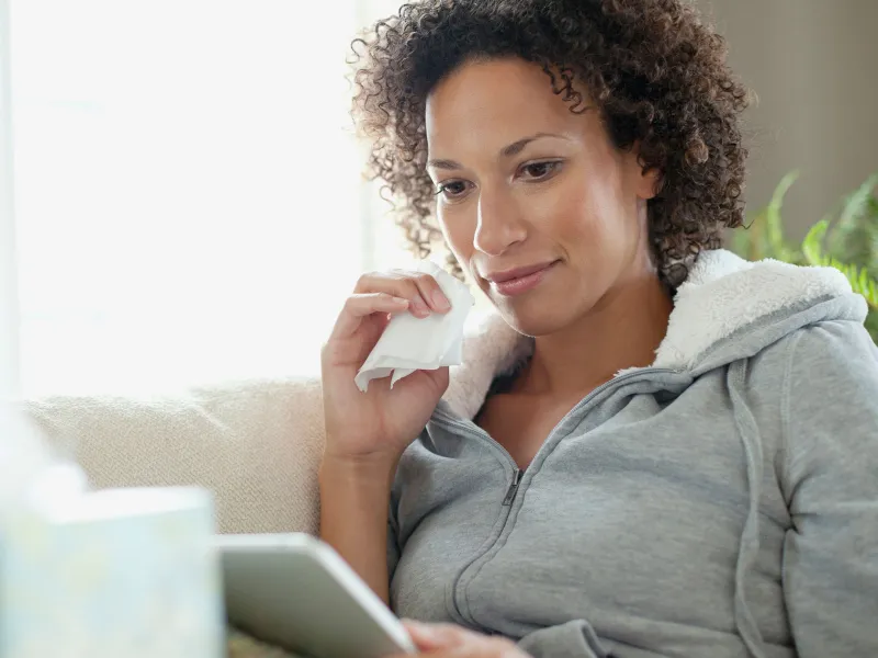 An African American woman dabs her face with a tissue while scoping out her tablet.