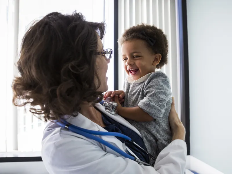 A female pediatrician holds a smiling infant in an exam room. 