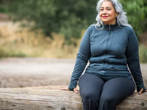 A mature woman sits outside on a log 