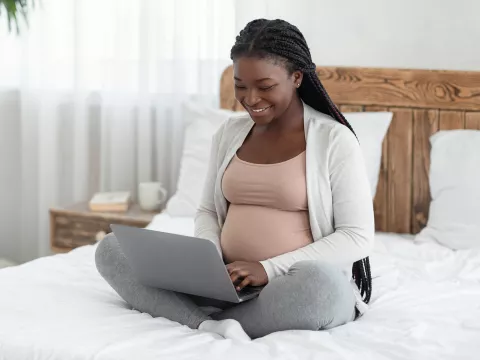 A Smiling Pregnant Woman Sits on Her Bed, in Her Home, Using a Laptop 