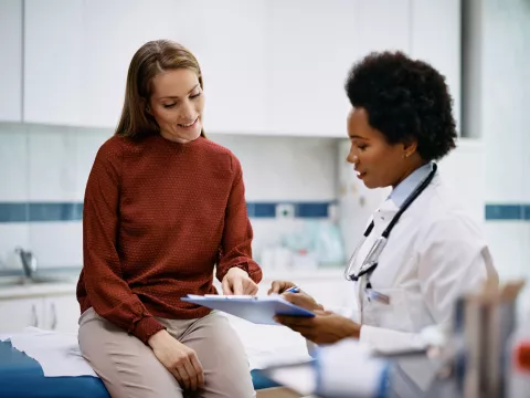 A Doctor Speaks to Her Patient in an Exam Room While Going Over Her Charts on a Laptop.