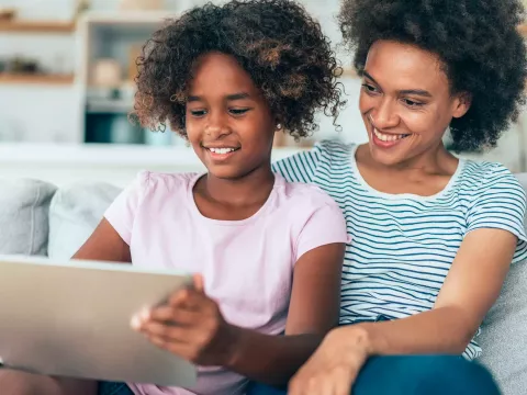 A MOther and Daughter Smile as They Sit Together and Surf the Internet on a Tablet.