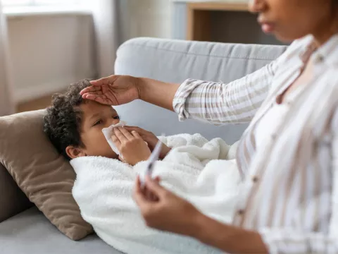 A Small Child Blows His Nose Laying on a Couch While His Mother Checks His Temperature