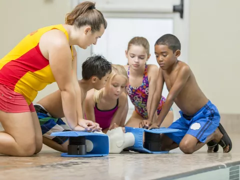Four children learn CPR from an adult poolside.