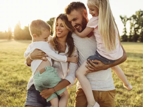 A family of four, the mother holding the son and the father holding the daughter, all laughing and smiling. The sun setting behind them.