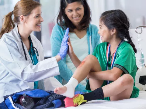 A child being treated for a sprained ankle at the ER, high fiving the ER doctor, while her mom looks on and smiles.