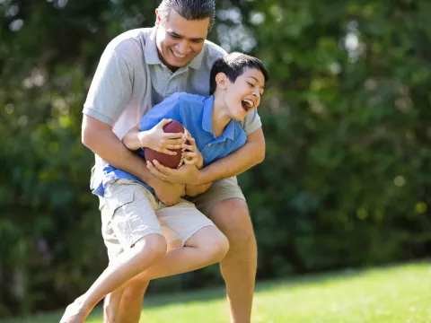 A father playfully tackles his son as they play football outside.