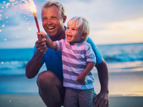 A father and son holding a firework on the beach at sunset.