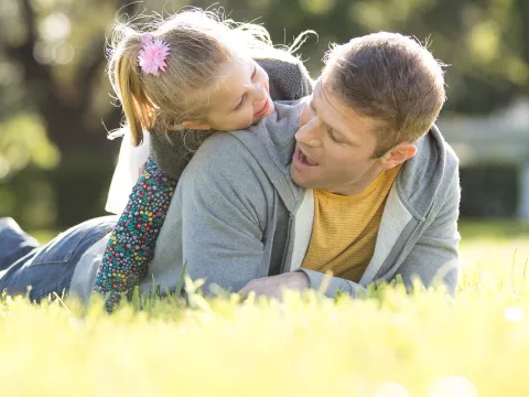 A little girl climbing on her fathers back laying down in the grass.
