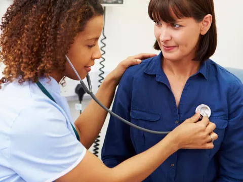 A woman sitting on an exam table, having her lungs listened to by a doctor.