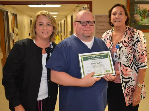 Three people standing in a hall holding a Daisy Award.