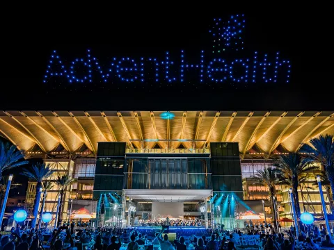 AdventHealth Orchestra performing at a night outdoor concert at Dr. Phillips Center for the Performing Arts with drones making the AdventHealth logo in the sky.