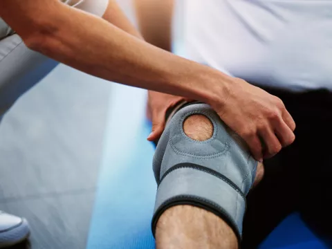 A man sitting down on a blue mat with a grey knee brace on, stretching his leg as another person helps him.