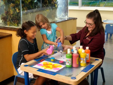 Two children and an adult woman sitting at a low arts and crafts table doing crafts.