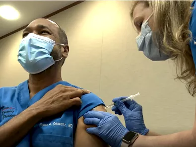 Dr. Schwartz rolls up his blue scrub sleeve as nurse administers the COVID-19 Vaccine