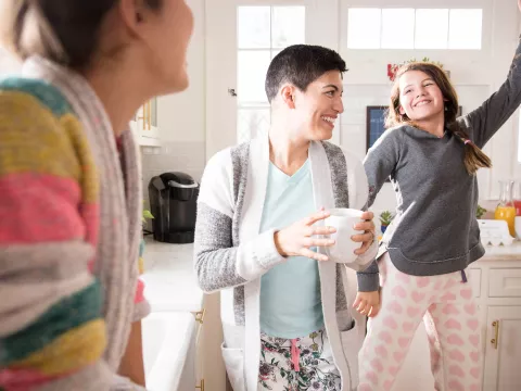 A woman dancing in the kitchen with her daughters and drinking coffee.
