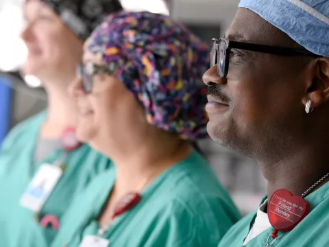 Three surgical nurses stand in the OR and look hopefully to the left of the image.