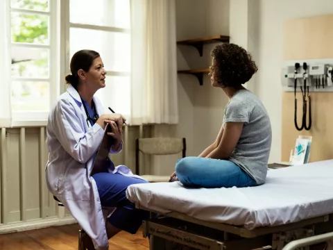 A young woman sits in a hospital room and talks about her health and wellness with her primary care doctor.