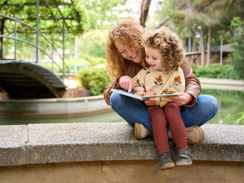 Mother and child reading a book together while sitting on a wall by a river.