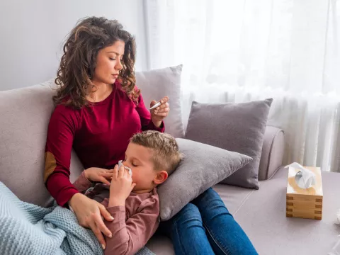 A mother and son on a couch. The mother is looking at a thermometer while the son lays his head on a pillow sitting on her lap while blowing his nose.